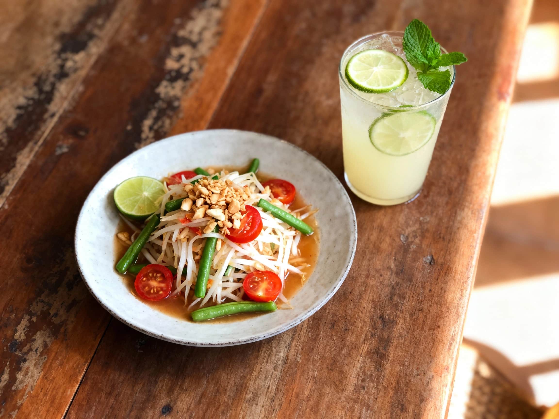 A vibrant Som Tum, or Thai green papaya salad, is presented in an rustic wooden bowl. Finely shredded green papaya forms the base, generously mixed with halved red cherry tomatoes and crunchy chopped roasted peanuts. A clear, zesty dressing glistens over the ingredients. Garnish includes fresh red chili slices and bright green lime wedges, enhancing the dish's visual appeal. The setting suggests a